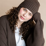 Modal wearing Chunky Knit Adult Ribbed Beanie in espresso over white t-shirt and chunky knit women's cardigan in espresso and blue jeans on a beige background