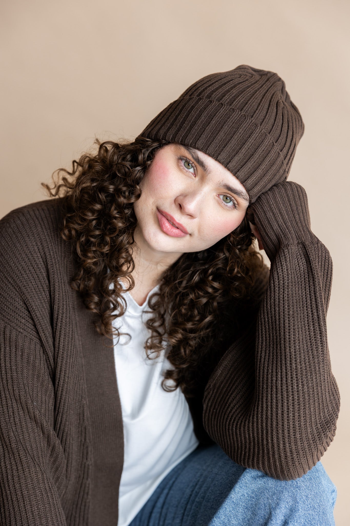 Modal wearing Chunky Knit Adult Ribbed Beanie in espresso over white t-shirt and chunky knit women's cardigan in espresso and blue jeans on a beige background
