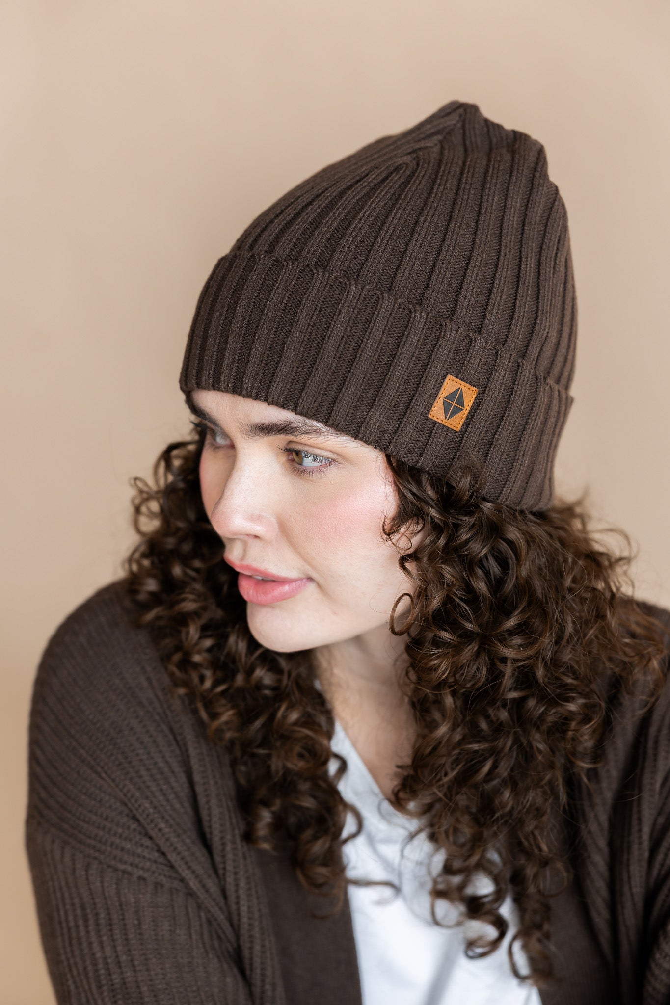 Modal wearing Chunky Knit Adult Ribbed Beanie in espresso over white t-shirt and chunky knit women's cardigan in espresso on a beige background