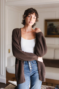 Modal standing in a living room wearing Chunky Knit Women's Waffle Cardigan in Espresso over white camisole and blue jeans
