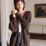 Modal standing in a living room wearing Chunky Knit Women's Waffle Cardigan in Espresso over white camisole and blue jeans