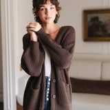 Modal standing in a living room wearing Chunky Knit Women's Waffle Cardigan in Espresso over white camisole and blue jeans