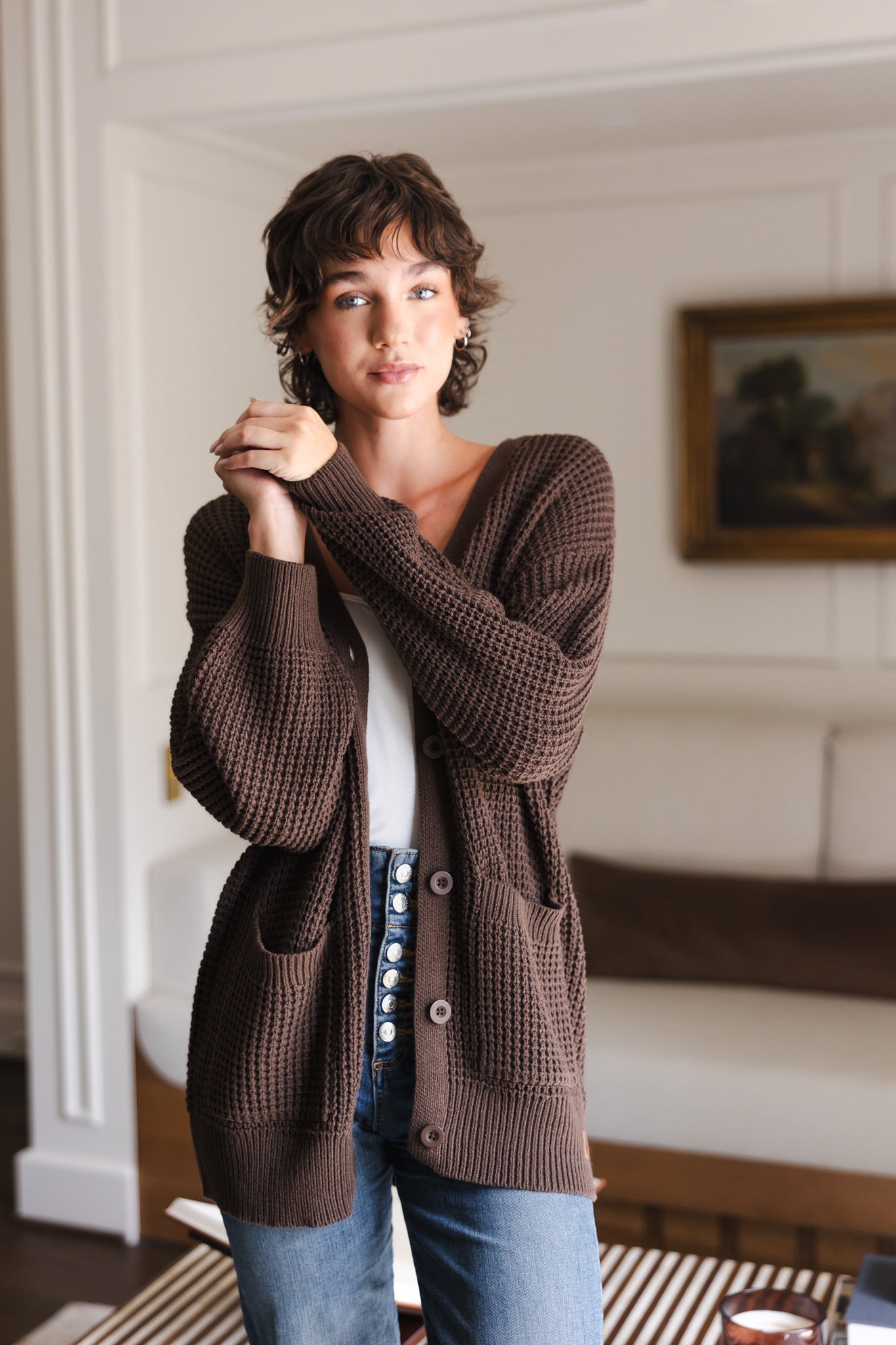 Modal standing in a living room wearing Chunky Knit Women's Waffle Cardigan in Espresso over white camisole and blue jeans