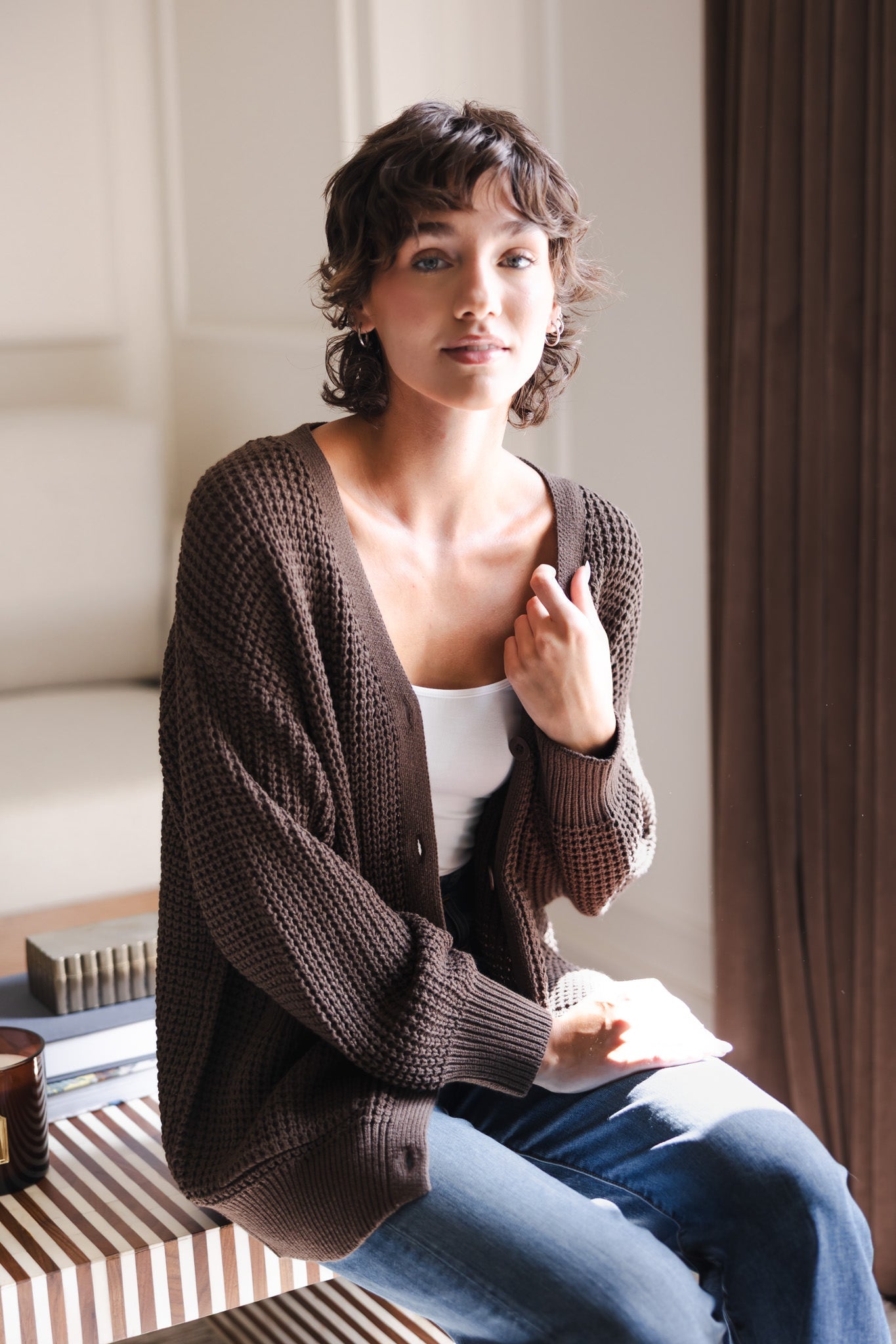Modal sitting in a living room wearing Chunky Knit Women's Waffle Cardigan in Espresso over white camisole and blue jeans