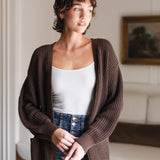 Modal posing in a living room wearing Chunky Knit Women's Waffle Cardigan in Espresso over white camisole and blue jeans