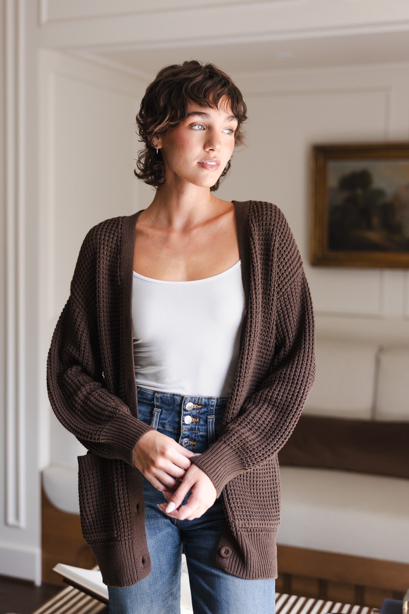 Modal posing in a living room wearing Chunky Knit Women's Waffle Cardigan in Espresso over white camisole and blue jeans