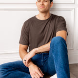 Man sitting in front of a white wall wearing Men's Crew Neck Tee in Espresso and blue jeans