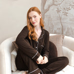 A woman with red hair sits cross-legged on a white armchair wearing a dark brown pajama set with white piping. The background features a beige wall with a framed bird painting and a gray tree-patterned wallpaper.