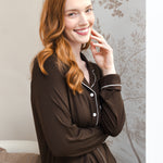 A smiling woman with red hair wearing a dark brown long-sleeve pajama set with white piping, posed in front of a patterned wallpaper background.