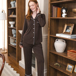 A woman wearing a dark brown pajama set with white piping stands barefoot in a cozy room with wooden bookshelves.