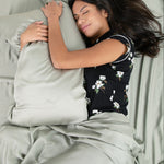 Female model laying in bed on the Bamboo Sateen Sheet Set in Eucalyptus holding a pillow wearing the short sleeve women's set in midnight big magnolia