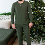 Smiling male standing beside a bed in front of a lit christmas tree wearing the Men's Ribbed Henley Set in Fir