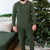 Smiling male standing beside a bed in front of a lit christmas tree wearing the Men's Ribbed Henley Set in Fir