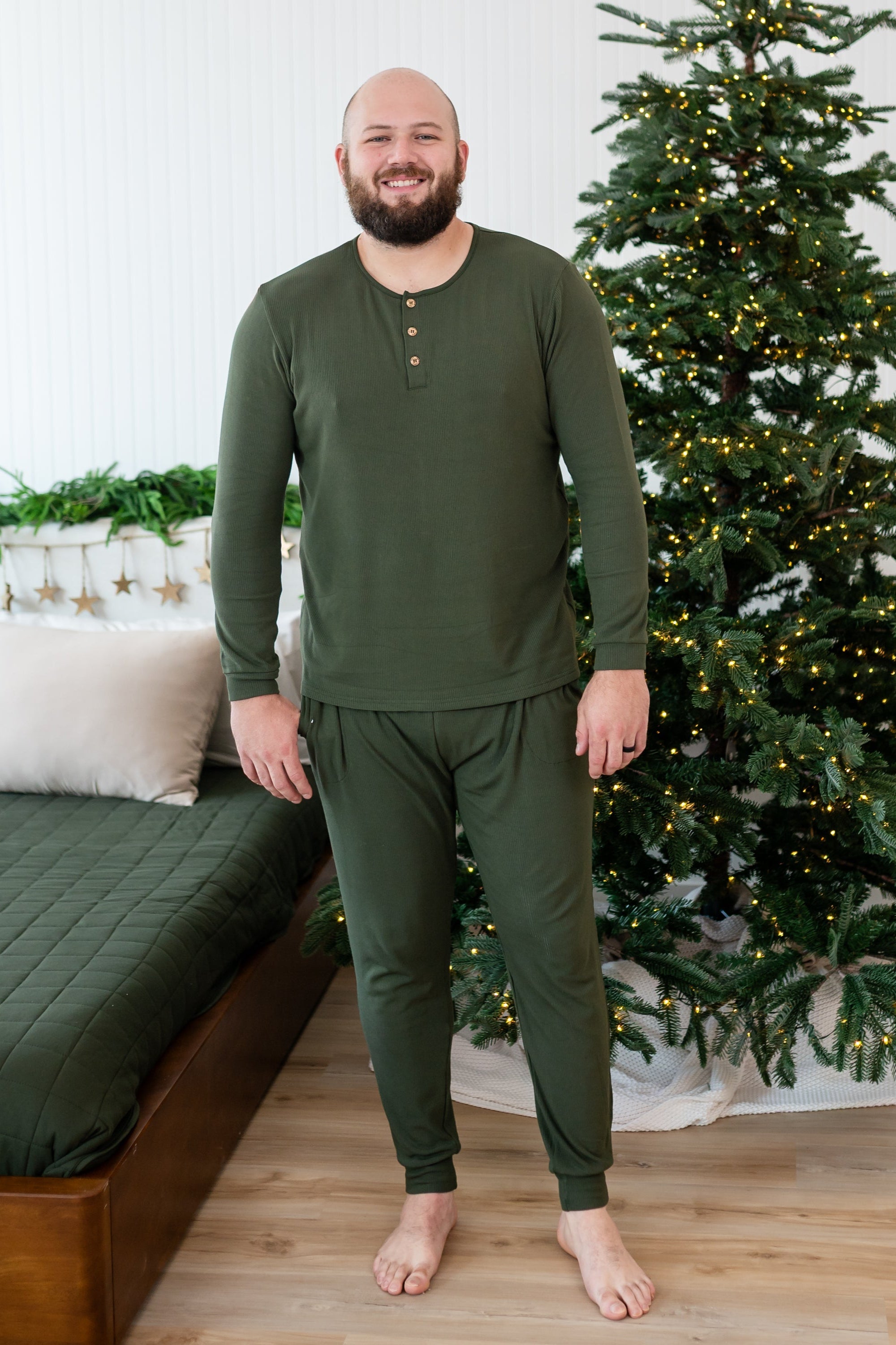 Smiling male standing beside a bed in front of a lit christmas tree wearing the Men's Ribbed Henley Set in Fir