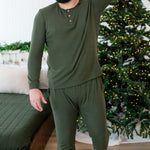 Male mode walking with one hand up behind his head wearing the Men's Ribbed Henley Set in Fir beside a bed and in front of a lit Christmas tree