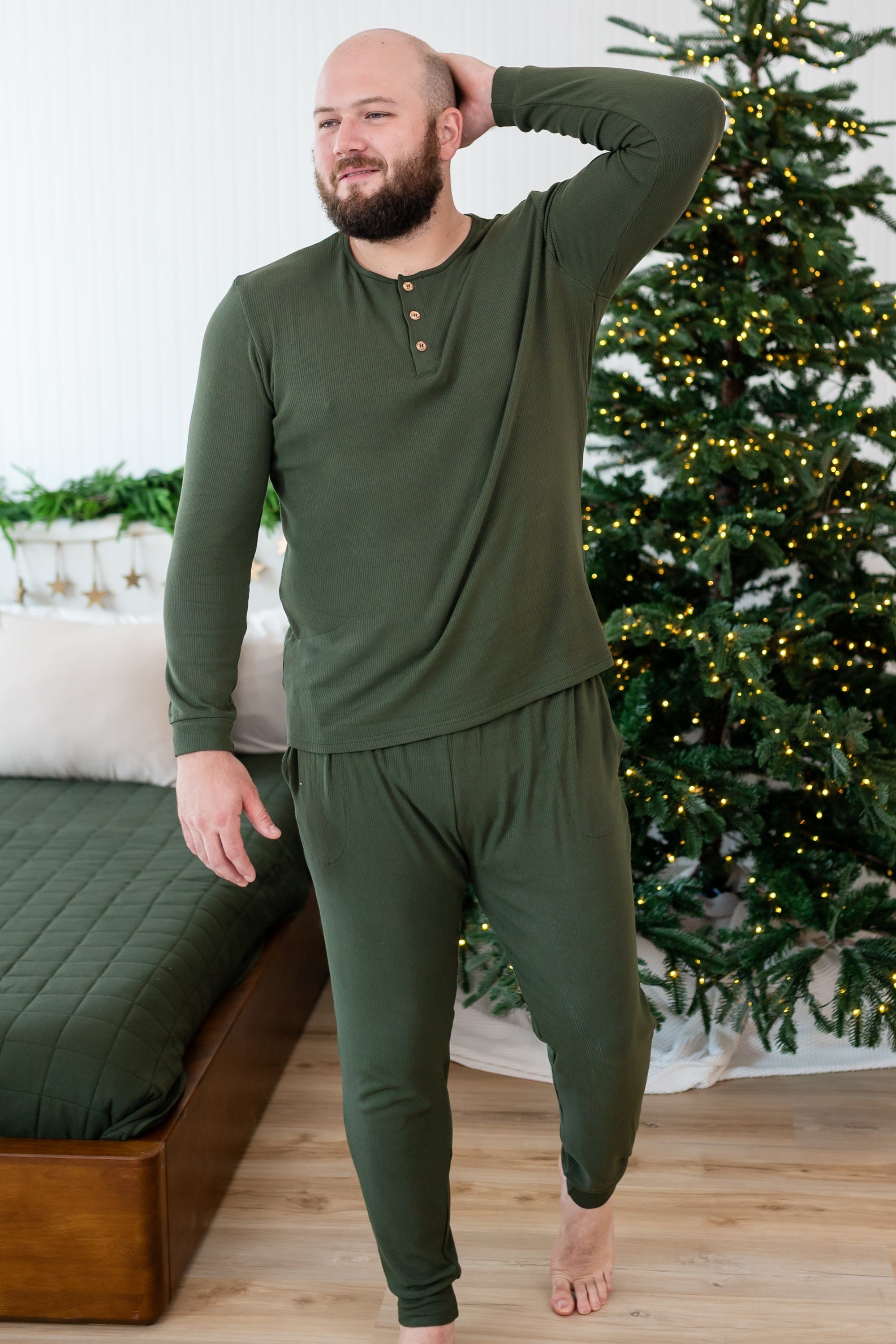 Male mode walking with one hand up behind his head wearing the Men's Ribbed Henley Set in Fir beside a bed and in front of a lit Christmas tree