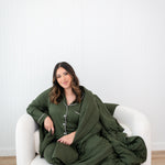 Model sitting on a white couch sofa wrapped in a the bamboo Quilt Blanket in Fir 2.5 and wearing the matching Long Sleeve Pajama Set