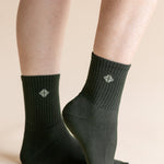 A pair of dark forest green crew socks worn on a person's feet, shown against a solid light beige background. The socks feature a ribbed cuff and a small white geometric logo on the front.