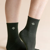 A pair of dark forest green crew socks worn on a person's feet, shown against a solid light beige background. The socks feature a ribbed cuff and a small white geometric logo on the front.
