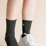 A close-up of a person's lower legs wearing a pair of dark charcoal gray ribbed crew socks and a white chunky sneaker against a solid beige background.