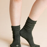 A close-up of a person's legs wearing dark olive green ribbed crew socks against a solid beige background.