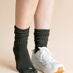 A person's legs wearing dark charcoal gray ribbed crew socks and a white athletic sneaker, set against a solid light beige background.