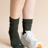 A person's legs wearing dark charcoal gray ribbed crew socks and a white athletic sneaker, set against a solid light beige background.