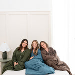 Smiling models sitting on a bed wearing Long Sleeve Pajama Set and wrapped in matching blanket in Fir, Atlantic and Espresso n