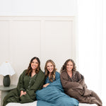 Models sitting on a bed while wrapped in Quilted Blankets 2.5 and wearing matching Long Sleeve Pajama Sets in Fir, Atlantic and Espresso 