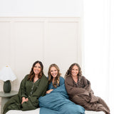 Models sitting on a bed while wrapped in Quilted Blankets 2.5 and wearing matching Long Sleeve Pajama Sets in Fir, Atlantic and Espresso 