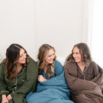 Models snuggled on a bed wrapped in Quilted Blankets in Fir, Atlantic and Espresso 