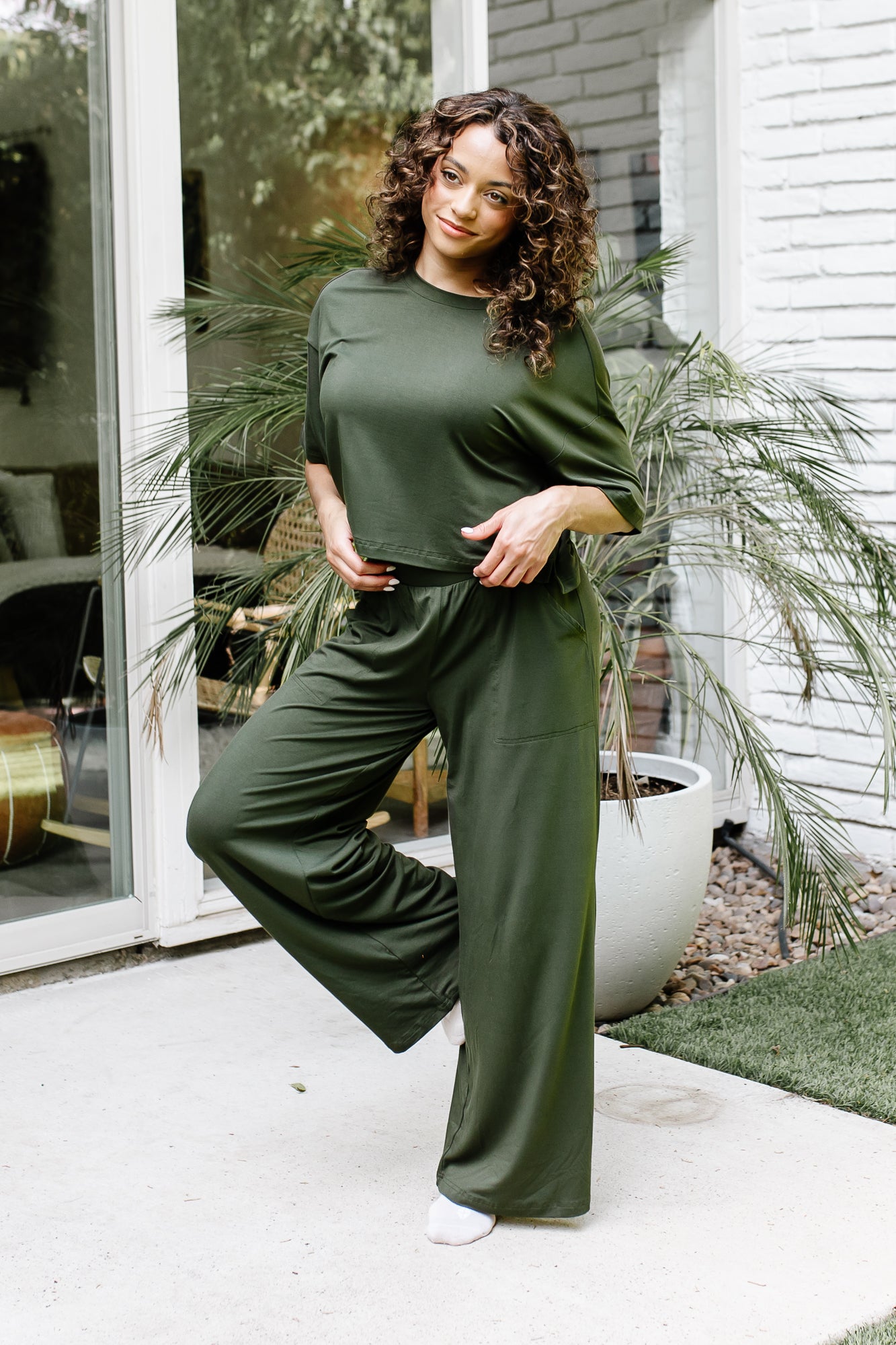 Female standing on a concrete patio in front of glass windows on a white brick house wearing the Women's Wide Leg Pant in Fir with matching short sleeve tee
