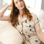 A woman with long red hair reclines on a white sofa wearing a short-sleeve pajama top and matching shorts. The pajamas are light beige with black piping and an allover print of small dog illustrations in black, orange, and tan.
