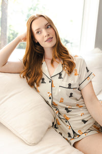 A woman with long red hair reclines on a white sofa wearing a short-sleeve pajama top and matching shorts. The pajamas are light beige with black piping and an allover print of small dog illustrations in black, orange, and tan.