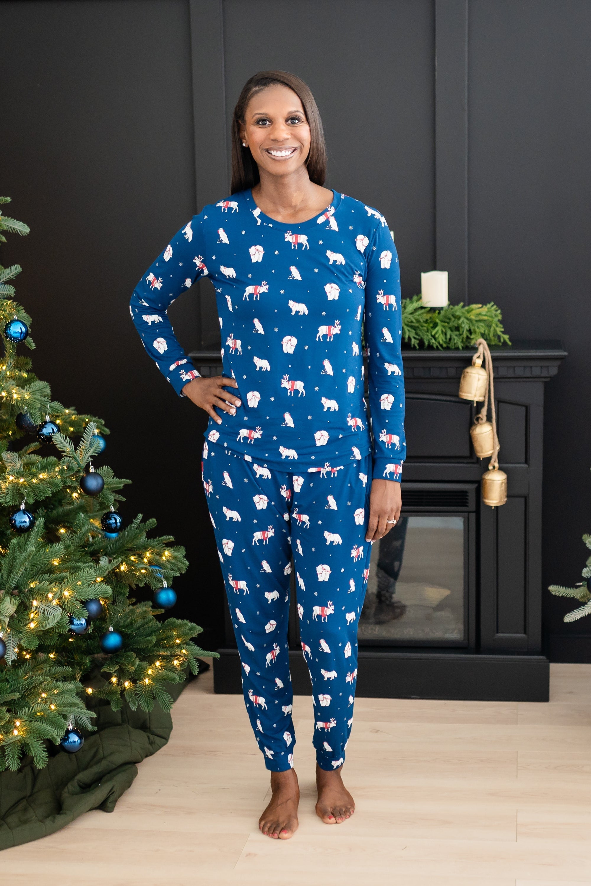 Smiling female model standing with one hand on her hip wearing the Women's Jogger Pajama Set in Frosty Friends in front of a black fireplace and between two decorated Christmas trees