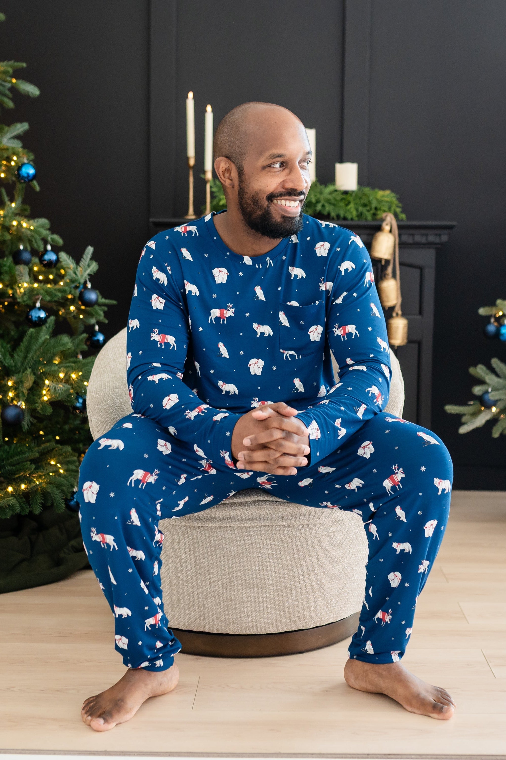 Male model sitting on a cushioned chair wearing the Men's Jogger Set in Frosty Friends in front of two decorated Christmas trees and a black fireplace