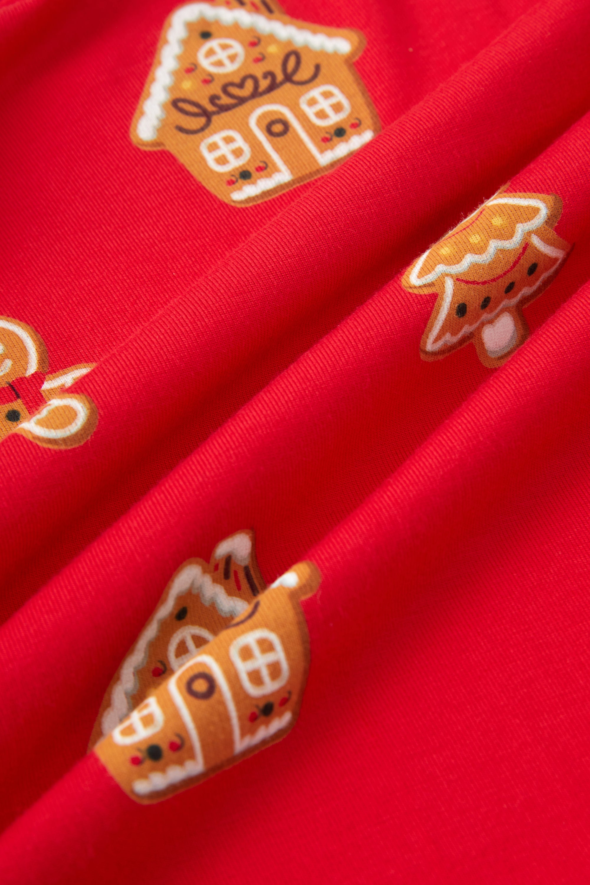 A close-up of bright red fabric with repeating gingerbread house and tree motifs in orange, white, and brown.
---1605 1