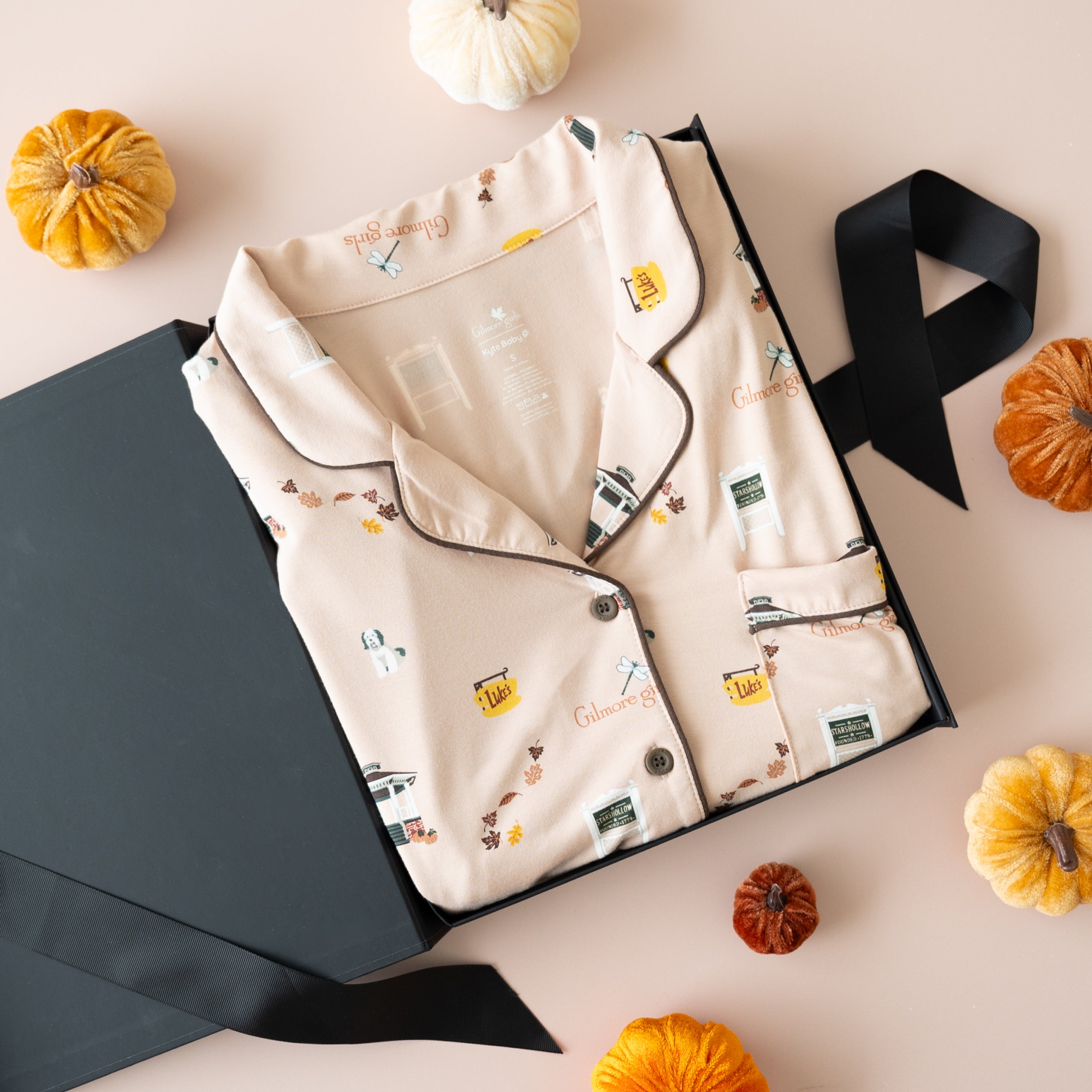 A flat-lay of a beige pajama set with a vintage gilmore girls-themed print, presented in a black gift box with a black ribbon, surrounded by small decorative pumpkins on a light pink background.