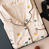 A flat-lay of a beige pajama set with a vintage gilmore girls-themed print, presented in a black gift box with a black ribbon, surrounded by small decorative pumpkins on a light pink background.