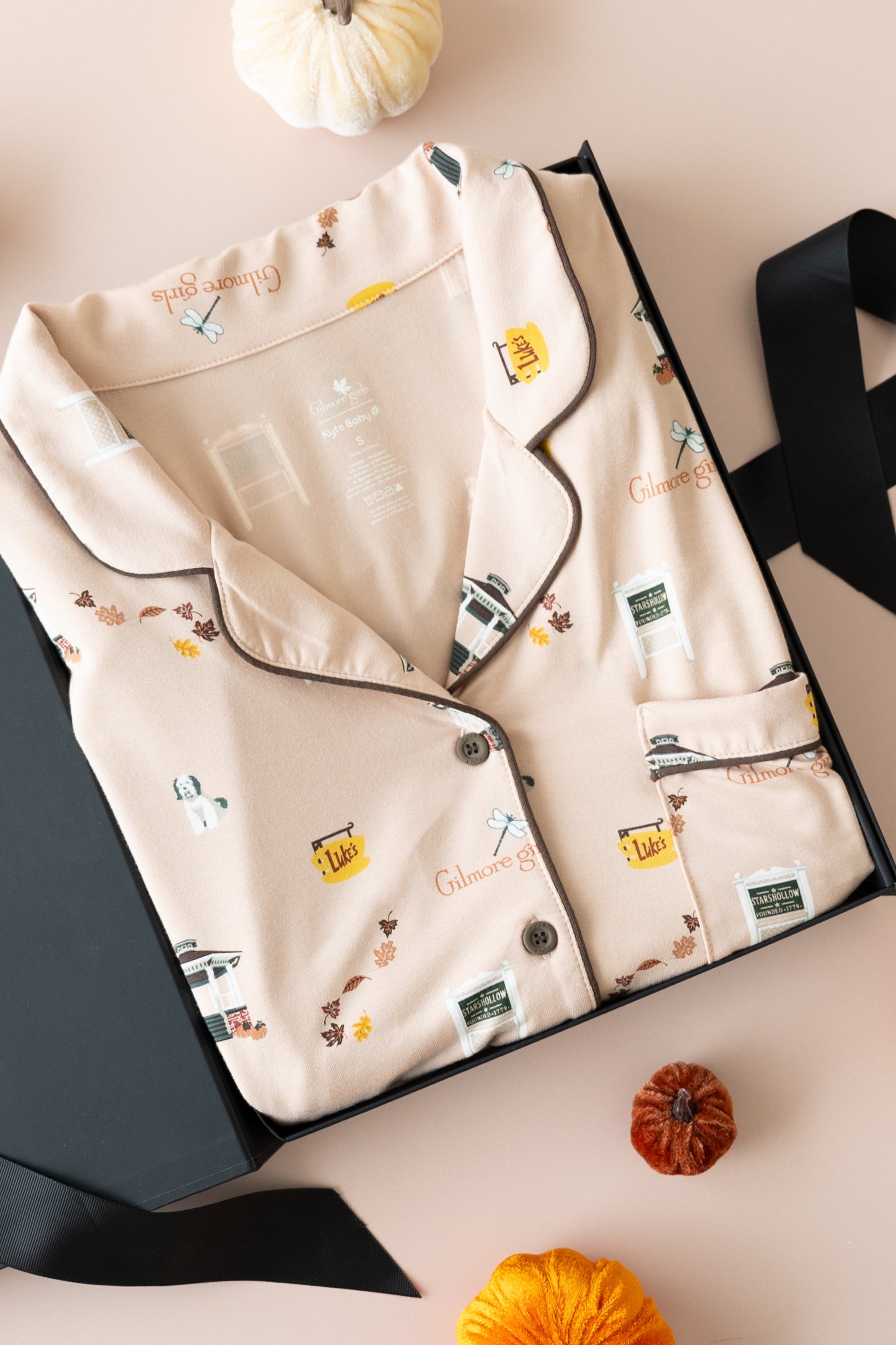 A flat-lay of a beige pajama set with a vintage gilmore girls-themed print, presented in a black gift box with a black ribbon, surrounded by small decorative pumpkins on a light pink background.
