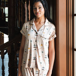 A woman wearing a short-sleeve pajama shirt and matching shorts with a small illustrated Gilmore Girls print, standing indoors near a wooden staircase and window. ---PJ 4