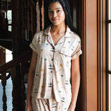 A woman wearing a short-sleeve pajama shirt and matching shorts with a small illustrated Gilmore Girls print, standing indoors near a wooden staircase and window. ---PJ 4