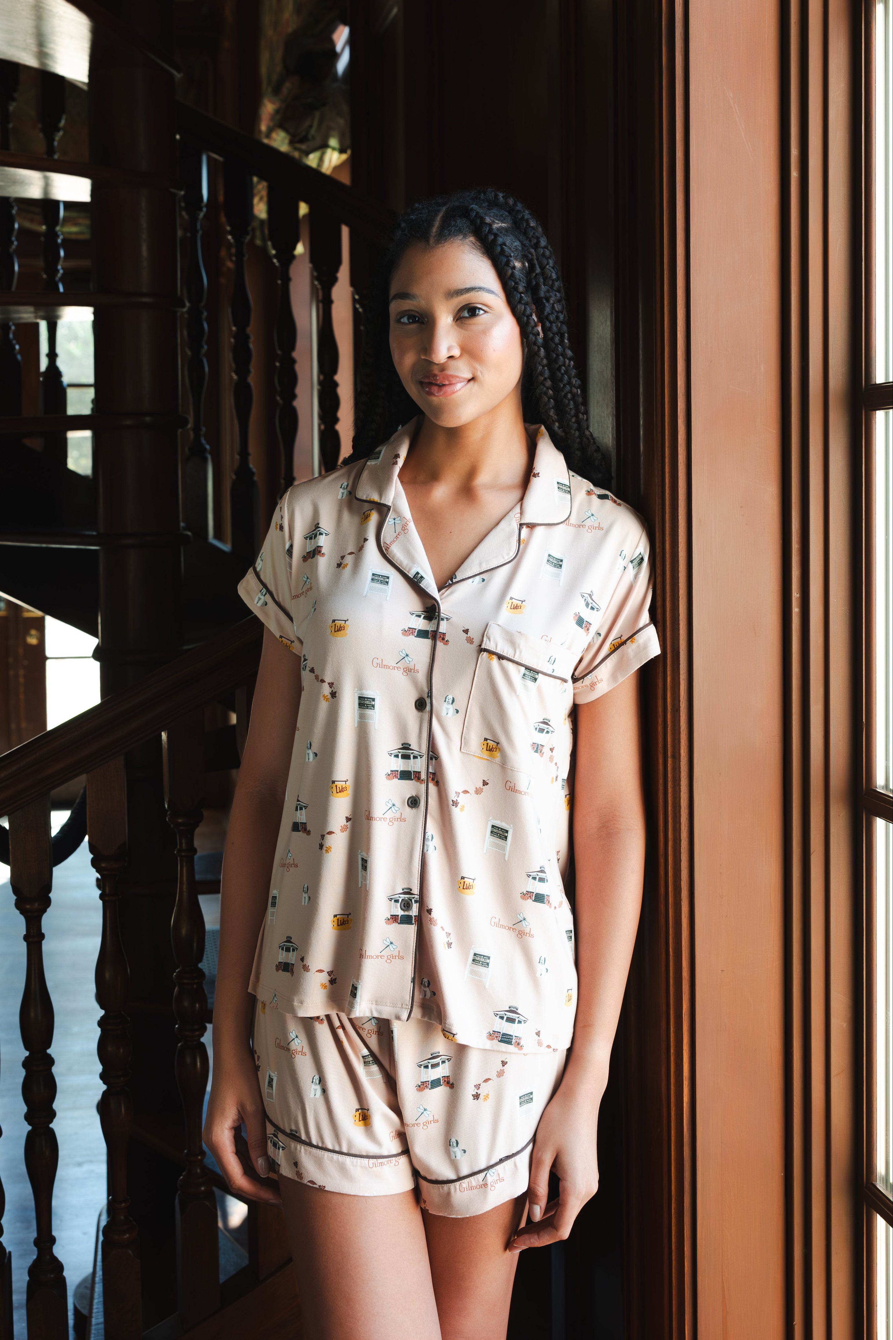 A woman wearing a short-sleeve pajama shirt and matching shorts with a small illustrated Gilmore Girls print, standing indoors near a wooden staircase and window. ---PJ 4