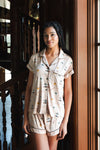 A woman wearing a short-sleeve pajama shirt and matching shorts with a small illustrated Gilmore Girls print, standing indoors near a wooden staircase and window. ---PJ 4