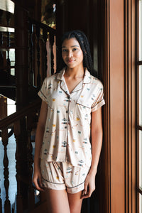 A woman wearing a short-sleeve pajama shirt and matching shorts with a small illustrated Gilmore Girls print, standing indoors near a wooden staircase and window. ---PJ 4