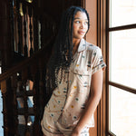 A woman wearing a beige short-sleeve pajama set with a small multicolor Gilmore Girls print stands beside a large window in a warm-toned interior with a wooden staircase.
