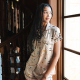 A woman wearing a beige short-sleeve pajama set with a small multicolor Gilmore Girls print stands beside a large window in a warm-toned interior with a wooden staircase.