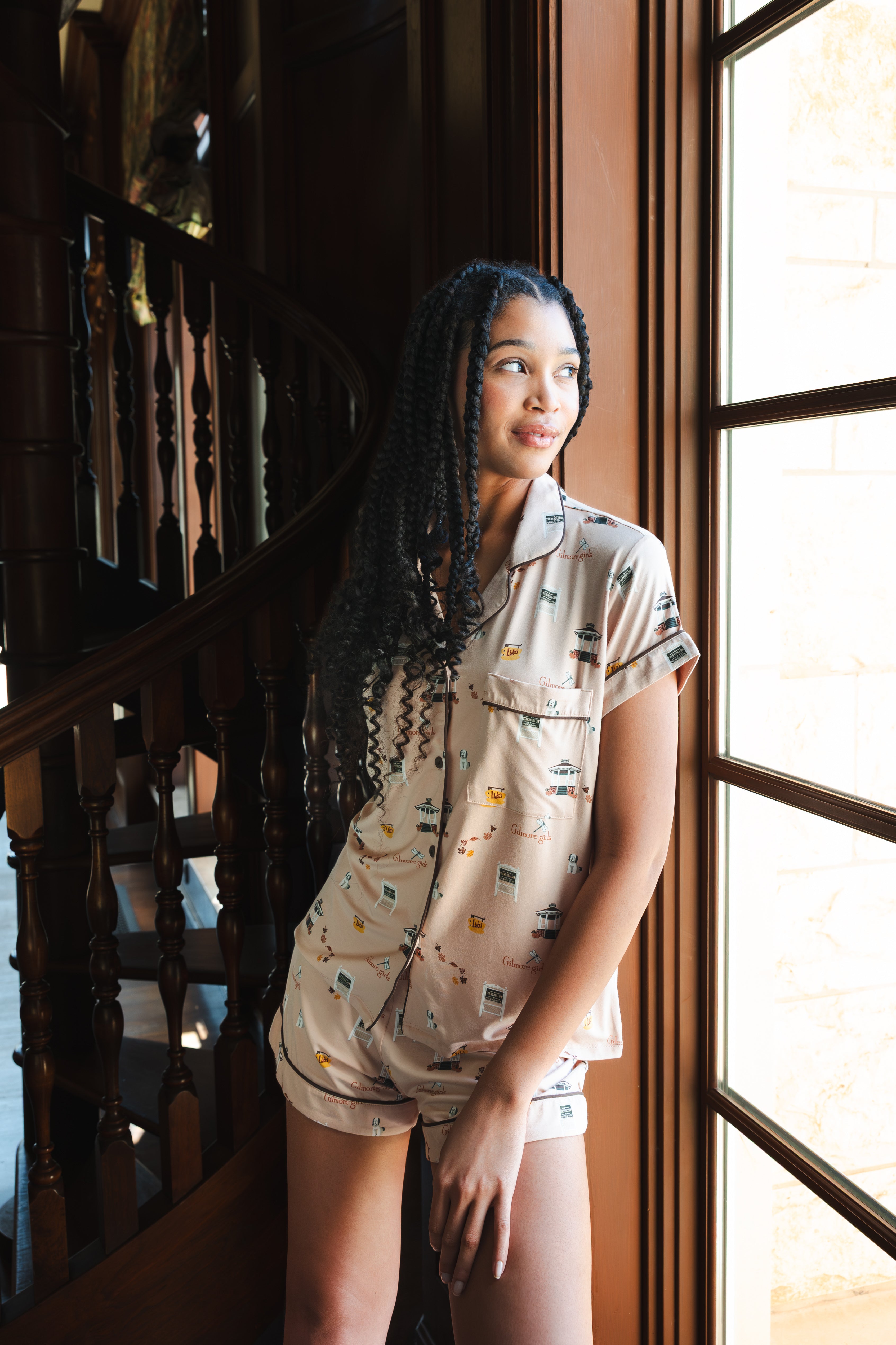 A woman wearing a beige short-sleeve pajama set with a small multicolor Gilmore Girls print stands beside a large window in a warm-toned interior with a wooden staircase.