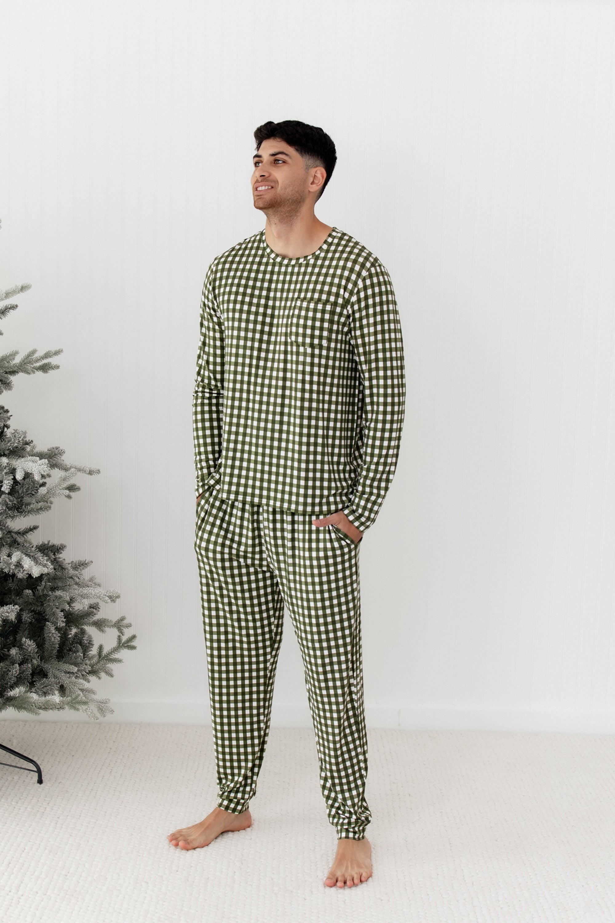 Man wearing green checkered pajamas standing in front of a white wall with a Christmas tree.