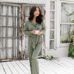 Model standing on a white wooden floor, wearing the Women's Long Sleeve Pajama Set in Gingham Fir holding a mug in front of white-paneled walls